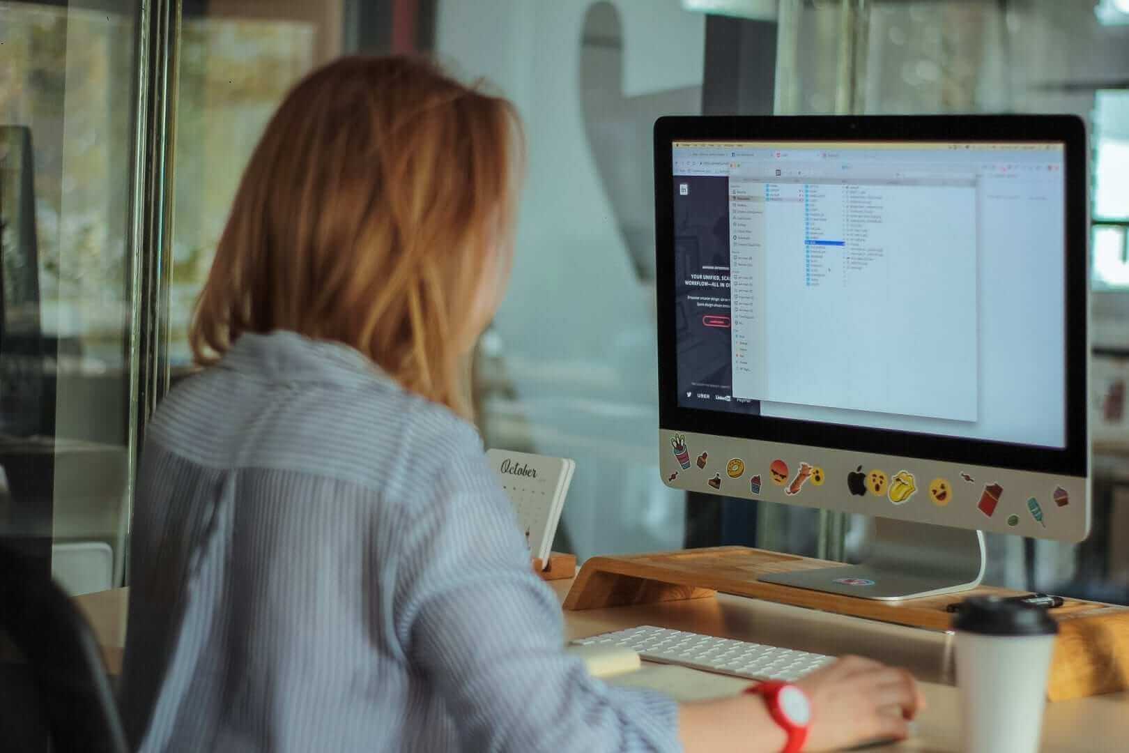 אדם עם שיער ארוך יושב ליד שולחן, משתמש ב-iMac מעוטר במדבקות. המסך מציג ספריית קבצים, אולי קשורה לפרויקטים של שירותי תרגום או לוקיזציה. כוס קפה טייק אווי מונחת בקרבת מקום במשרד.